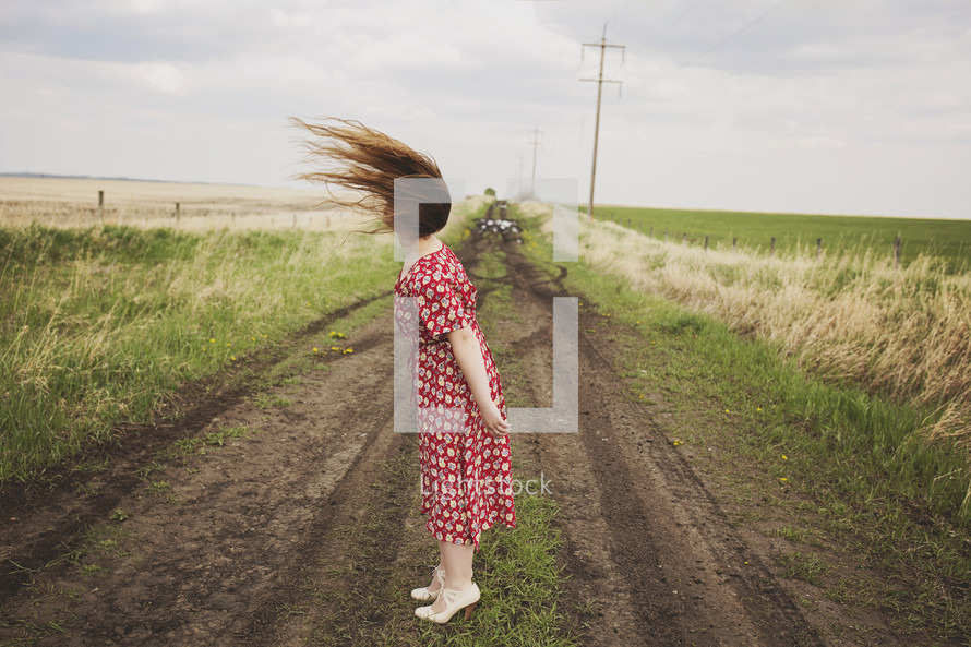 a woman standing alone in the middle of a dirt road with hair blowing 