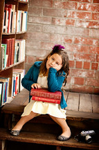 child sitting in a library with a stack of books in her lap and her head resting in her hand