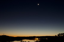 The last bit of sun sets below the horizon to reveal the night sky with moon overhead. A bridge can be seen in the distance.