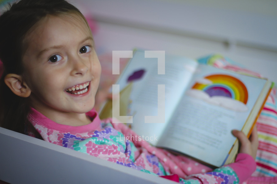 Girl in pajamas reading the Bible and smiling. She's reading the story of Noah before bedtime.  Reading a children's Bible.