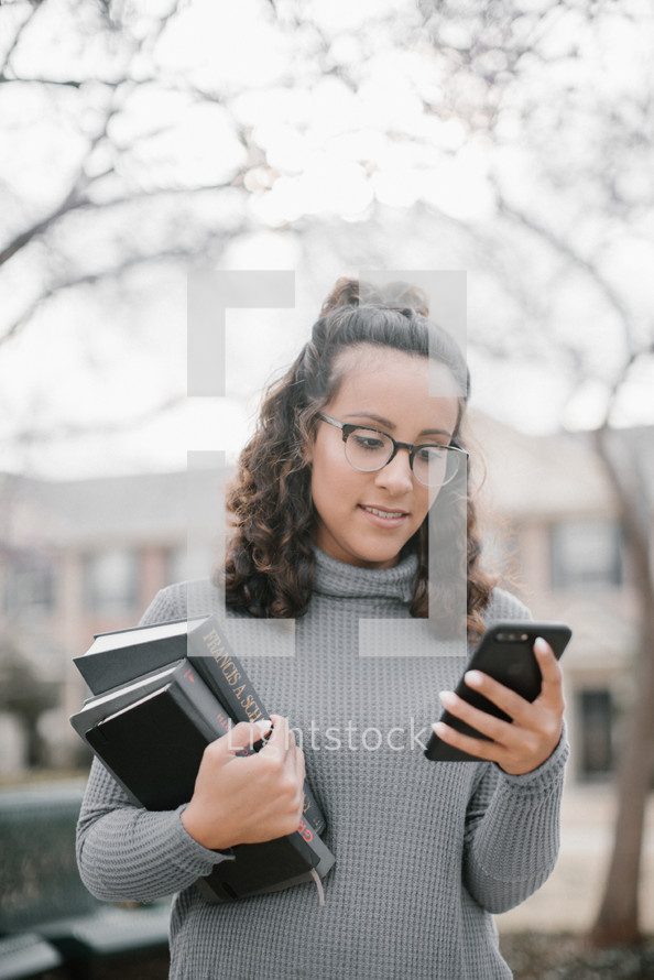 a young woman holding a stack of books and looking at her cellphone 