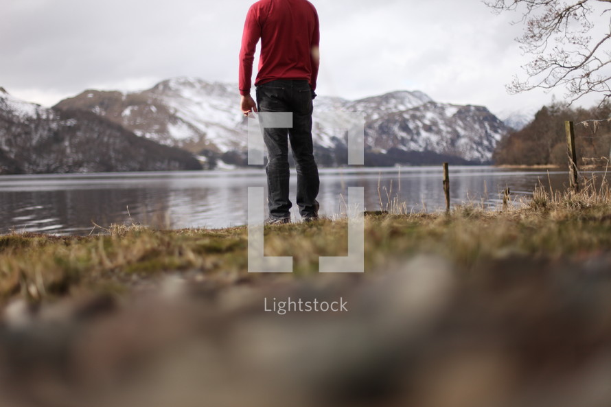 A man in a red shirt standing by a lake near a mountain range.