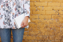 a woman standing in front of a yellow brick wall holding a Bible 
