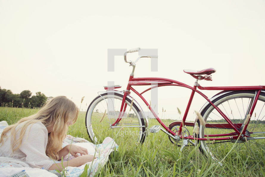young woman reading on a blanket in the grass, next to her bicycle 