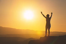 a woman with raised hands standing at the edge of a mountain at sunset 