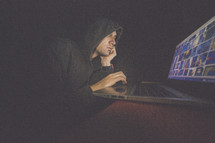 A man in a hooded sweatshirt works on a laptop computer in the dark.