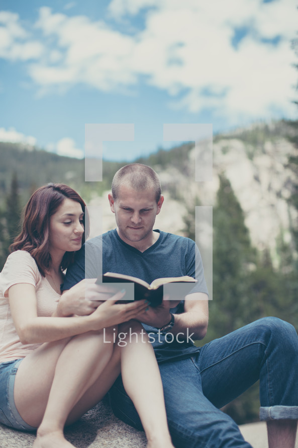 woman and man sitting on a rock reading a Bible by a river 