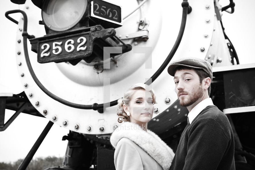 Couple, dressed in 1940's clothing, standing in front of the engine of a old train