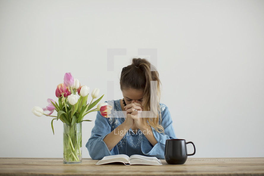 a woman sitting at a desk with a mug of coffee and a vase of tulips praying over a Bible 