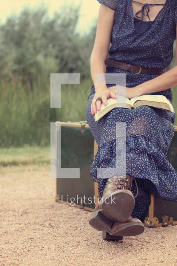 woman sitting on luggage on a dirt road reading a Bible 