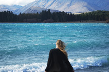 a woman standing on a shore looking out at the water and mountains 