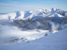 Mountain chalet in winter landscape with low altitude clouds in bright sunny day. Snow covered mountains