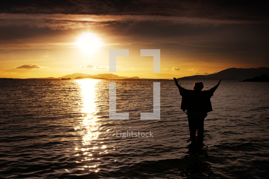 Man standing in ocean with arms raised at sunset.