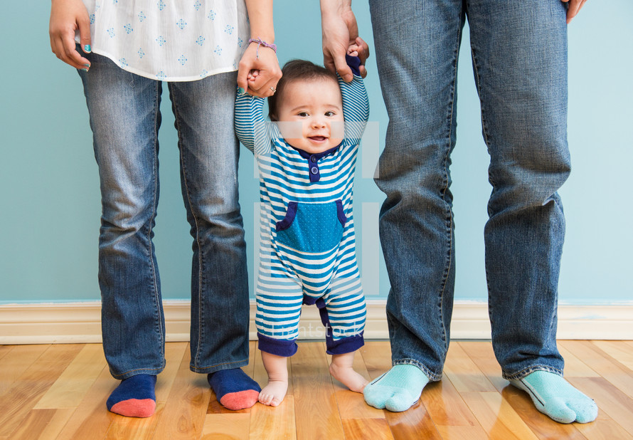 Infant child standing on the wood floor holding his parents' hands.