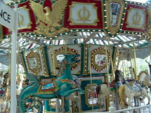 A colorful merry go round with a little blonde haired girl riding a merry go round with toy mechanical horses.
