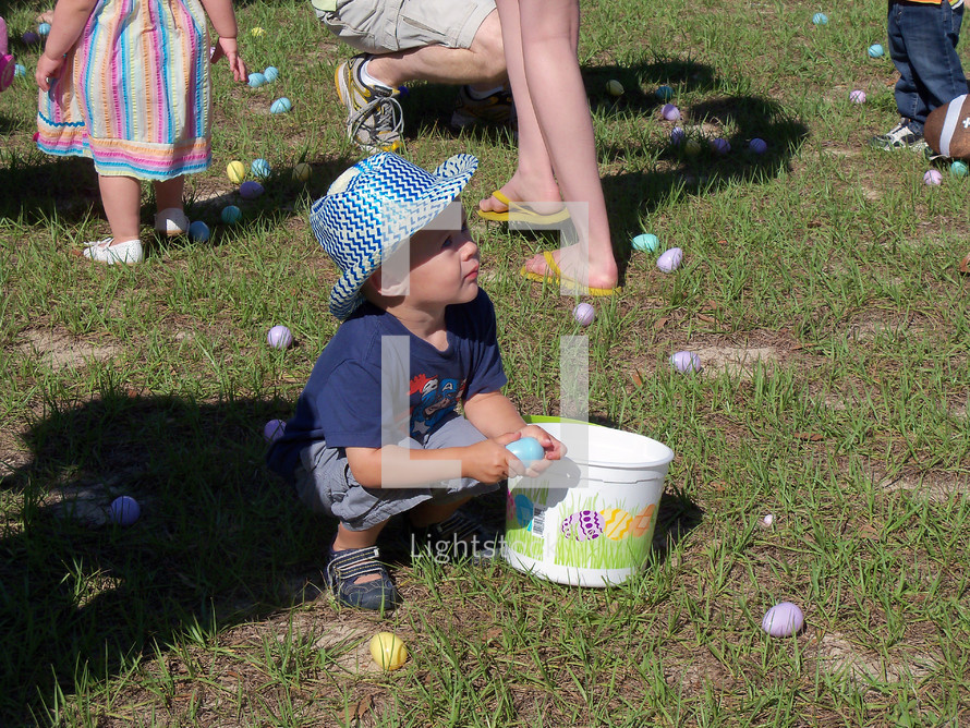 A young boy wearing a white and blue hat kneels down to pick up Easter Eggs among his friends and family at a church sponsored Easter Egg Hunt for children. Each year many churches gather the children and families of the community to have a church wide Easter Egg hunt and invite people to church. 