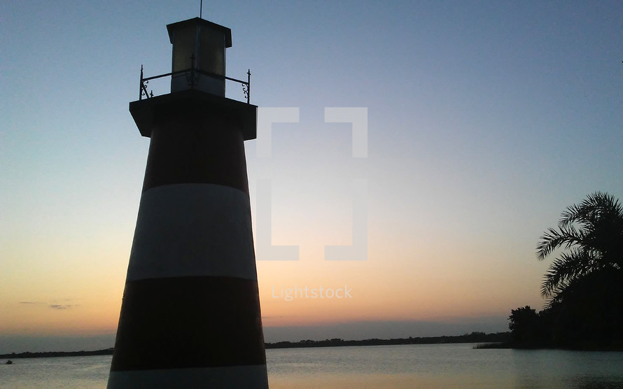 A lighthouse at dusk surrounded by calm water as the sun sets an orange glow along the horizon at dusk. 