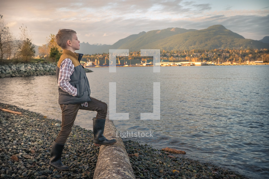 Young boy child in boots standing on a rocky shore thinking about his future. 