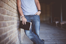 a man leaning against a brick wall holding a Bible 