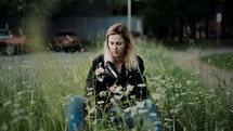 a young woman sitting in tall grass holding a camera 