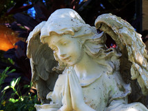 A statue of a praying female angel figure with a smiling face and prayerful pose adorns a courtyard in a church cemetery. 