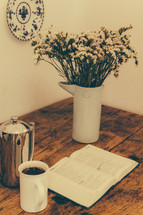 creamer, coffee mug, flowers in a vase and a Bible on a wood table 