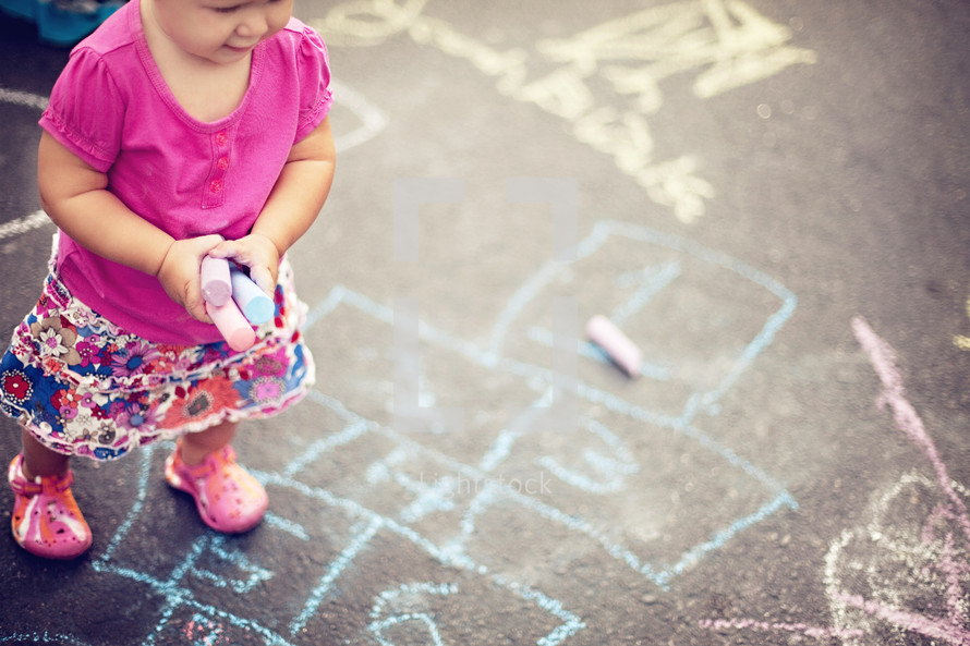 Aerial view of toddler girl in pink shirt with sidewalk chalk in her hands standing on sidewalk with chalk drawings on it.