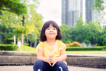 a toddler girl sitting in a park in a city 