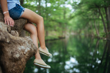 A girl in shorts dangles her legs from a tree over a river.