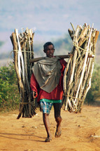 man walking bare foot on a dirt road carrying bundles of sticks 