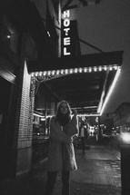 a woman standing on a city street under a hotel sign at night 