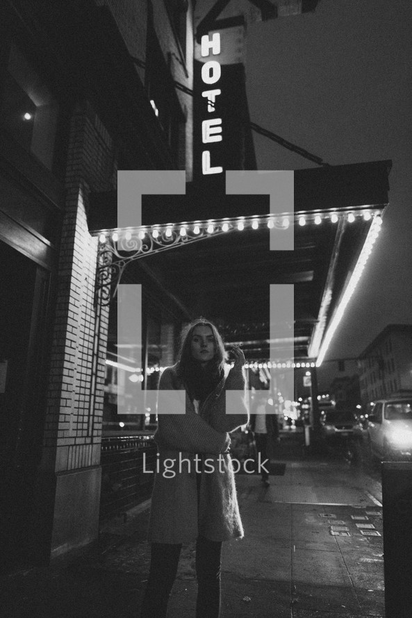 a woman standing on a city street under a hotel sign at night 