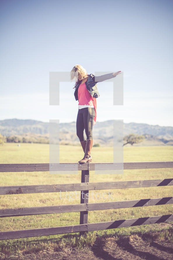 Woman balancing on a fence rail in a pasture with mountains.