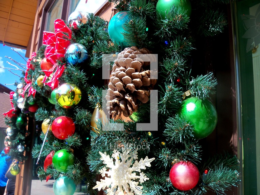 Christmas decorations adorning a holiday window wreath with snowflakes, red and green ornaments and a green evergreen adorned with red ribbons, pine cones and lively Christmas tree ornaments. 