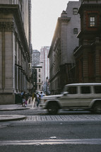 people on the sidewalk near an alley and passing vehicles 