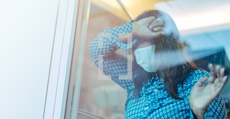 a sick woman wearing a face mask standing in a window 