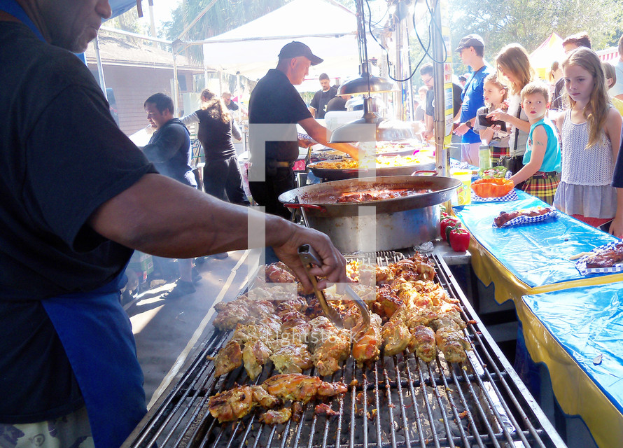 A group of Food vendors and cooks prepare a meal under a tent outdoors while young kids and adults wait for their food to be cooked and prepared at a Sunday brunch after church. 