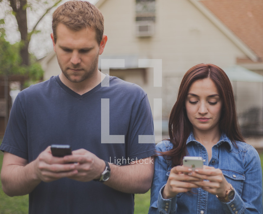couple standing next to each other texting on a cellphone 