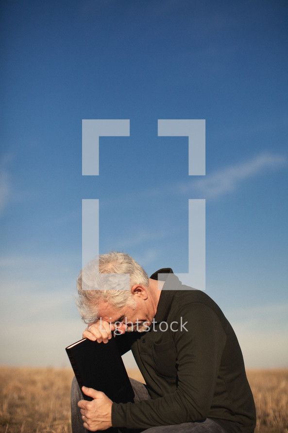 elderly man squatting in a field with his head bowed over a Bible in prayer