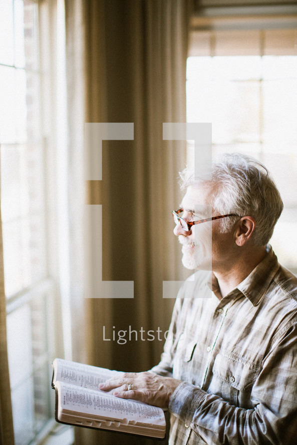 elderly man standing at a window reading a Bible by sunlight