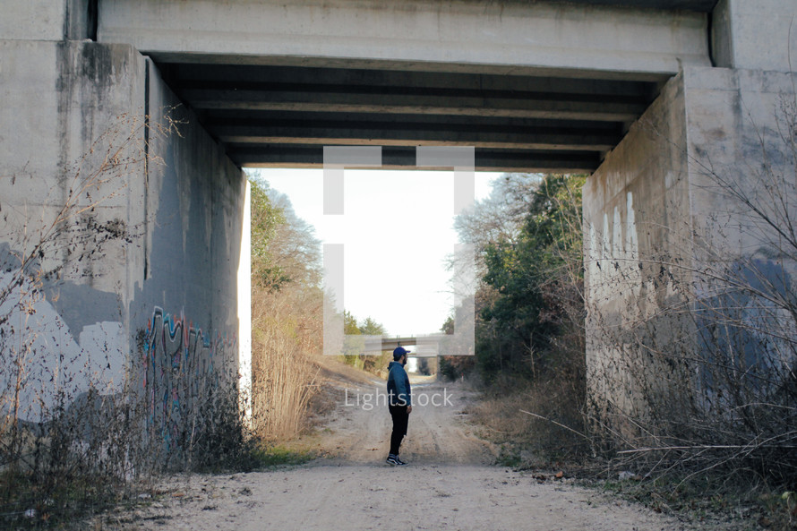man standing on a dirt road under a concrete overpass 