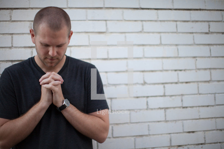 a man with praying hands and head bowed standing in front of a white brick wall 