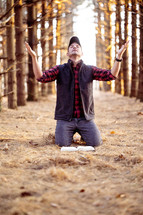 a man kneeling in a forest with hands raised 