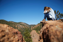 a woman sitting at the edge of a mountainside covering her face 
