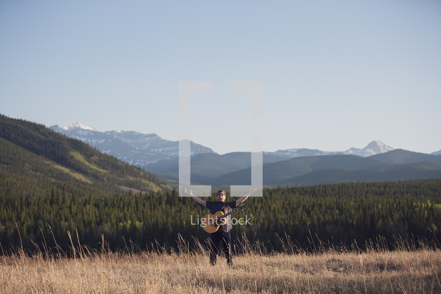 a man with a guitar standing in a field with raised arms and a mountain view 