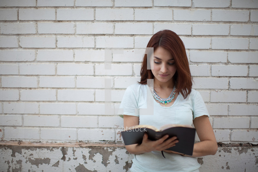 a young woman smiling as she reads a Bible in front of a white brick wall 