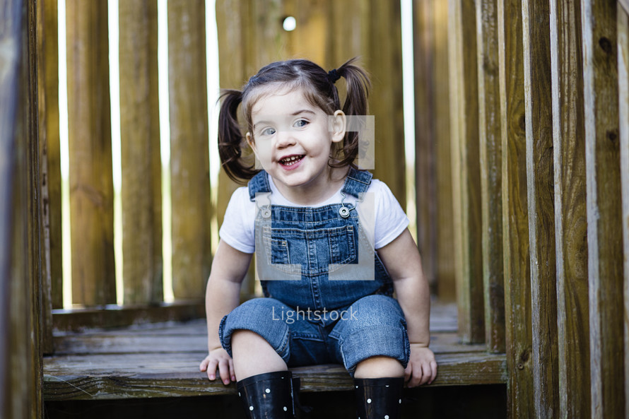 A little girl sitting on wooden steps in overalls.