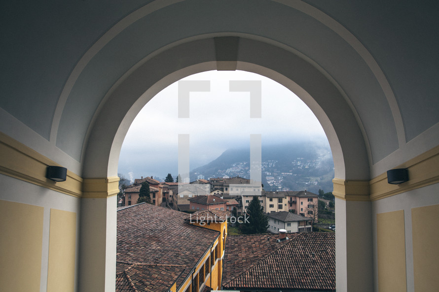 View through an archway of tile roofs, stucco buildings, and foggy mountains.