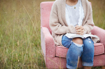 a woman sitting in a chair in a field with praying hands over the pages of a Bible 