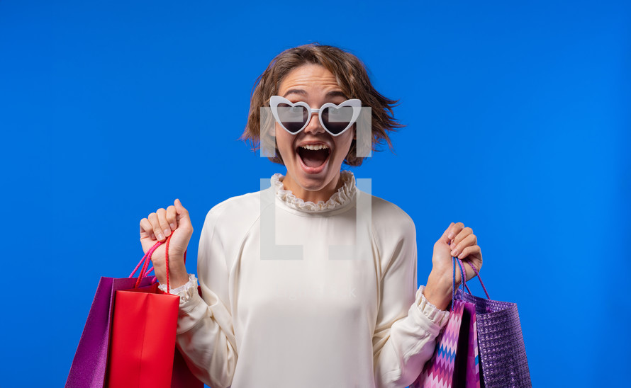 Excited woman with colorful paper bags after shopping on blue studio background. Concept of seasonal sale, purchases, spending money on gifts
